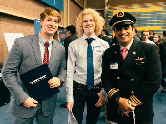Three people posing together at an indoor event, with two dressed in business attire and one in an airline pilot uniform, holding folders and smiling for the photo.