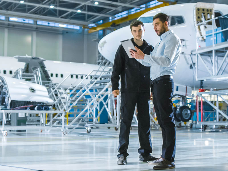Two professionals standing in an aircraft hangar reviewing information on a tablet, with aircraft and maintenance equipment in the background.