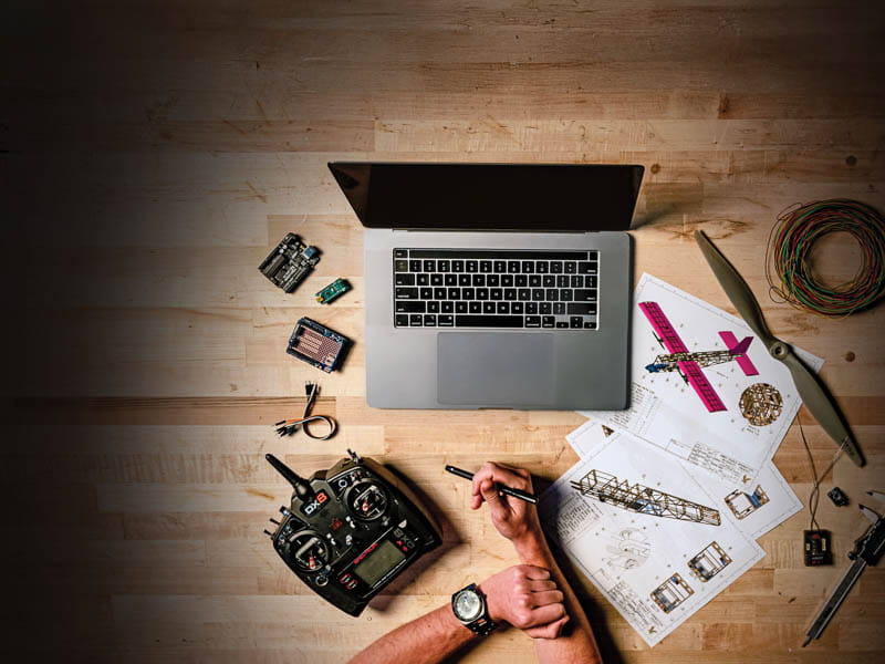 A desk with a laptop and drawings of planes, with a person taking notes, view from overhead.