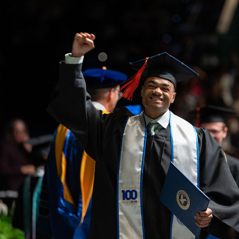 A man walks across the stage at a graduation ceremony, raising his arm in celebration after getting his diploma