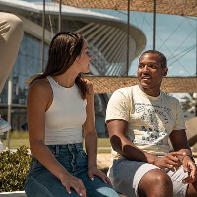 Two students seated outdoors, engaged in conversation, with a building in the background.