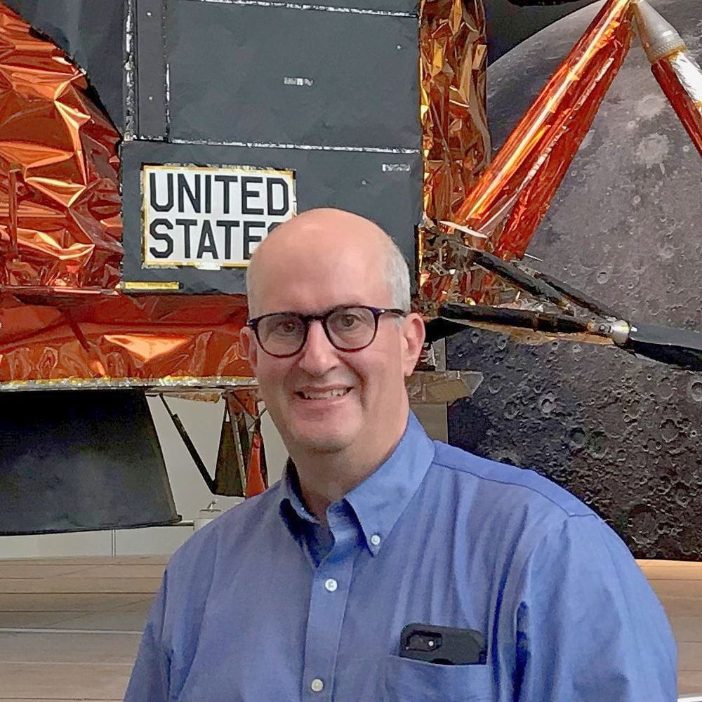 Sklar stands in front of an Apollo lunar module display with gold thermal foil, truss structure, and a ‘United States’ marking, Moon surface backdrop behind him.