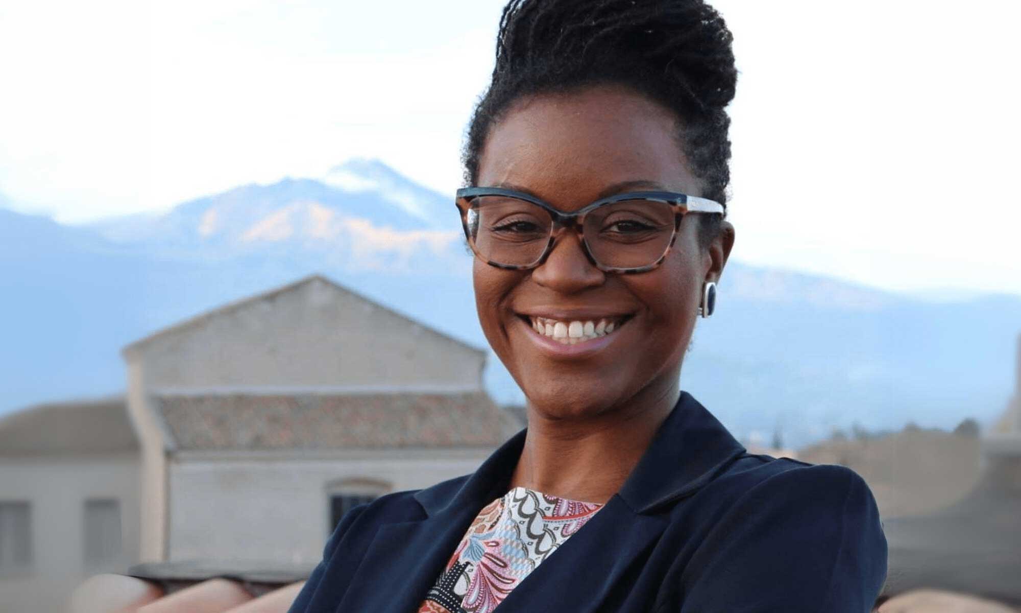 Person in a navy blazer smiles for a portrait, a home behind her and a mountain in the distance.