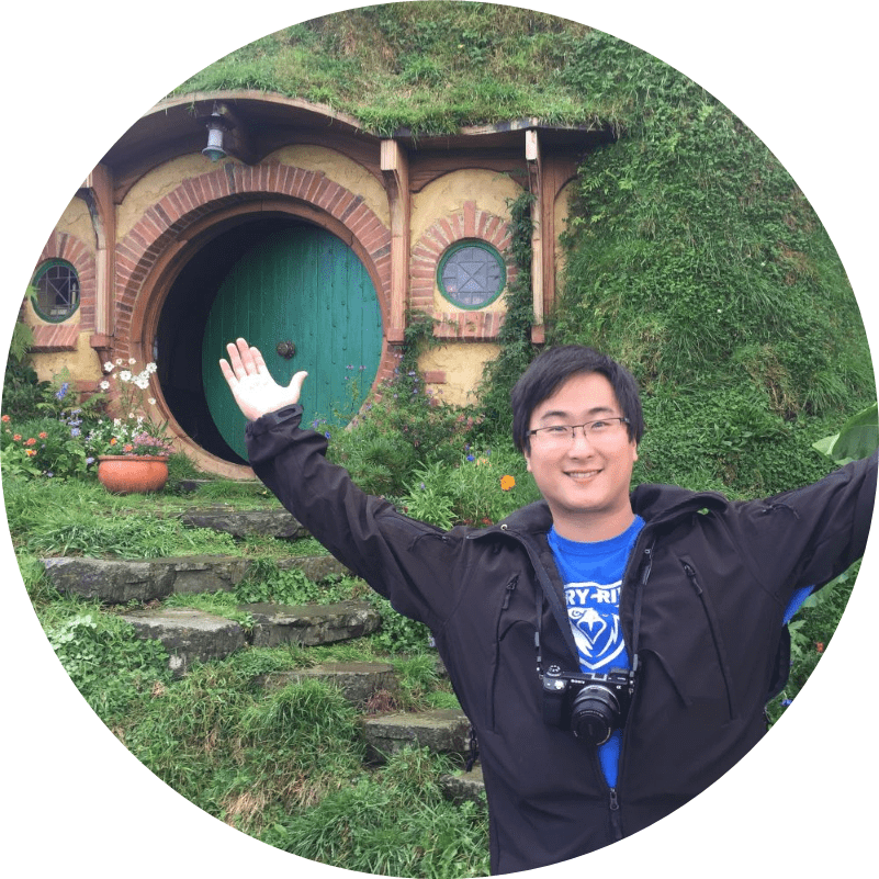 Christopher Awes in New Zealand posing with his arms outstretched in front of the round door of a hobbit house from the movie set.