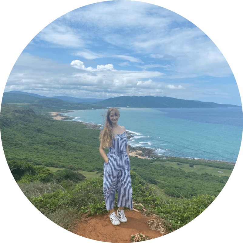 Carolyn Chatman standing on a cliff in Taiwan, overlooking a coastline with green hills and blue ocean under a partly cloudy sky. 