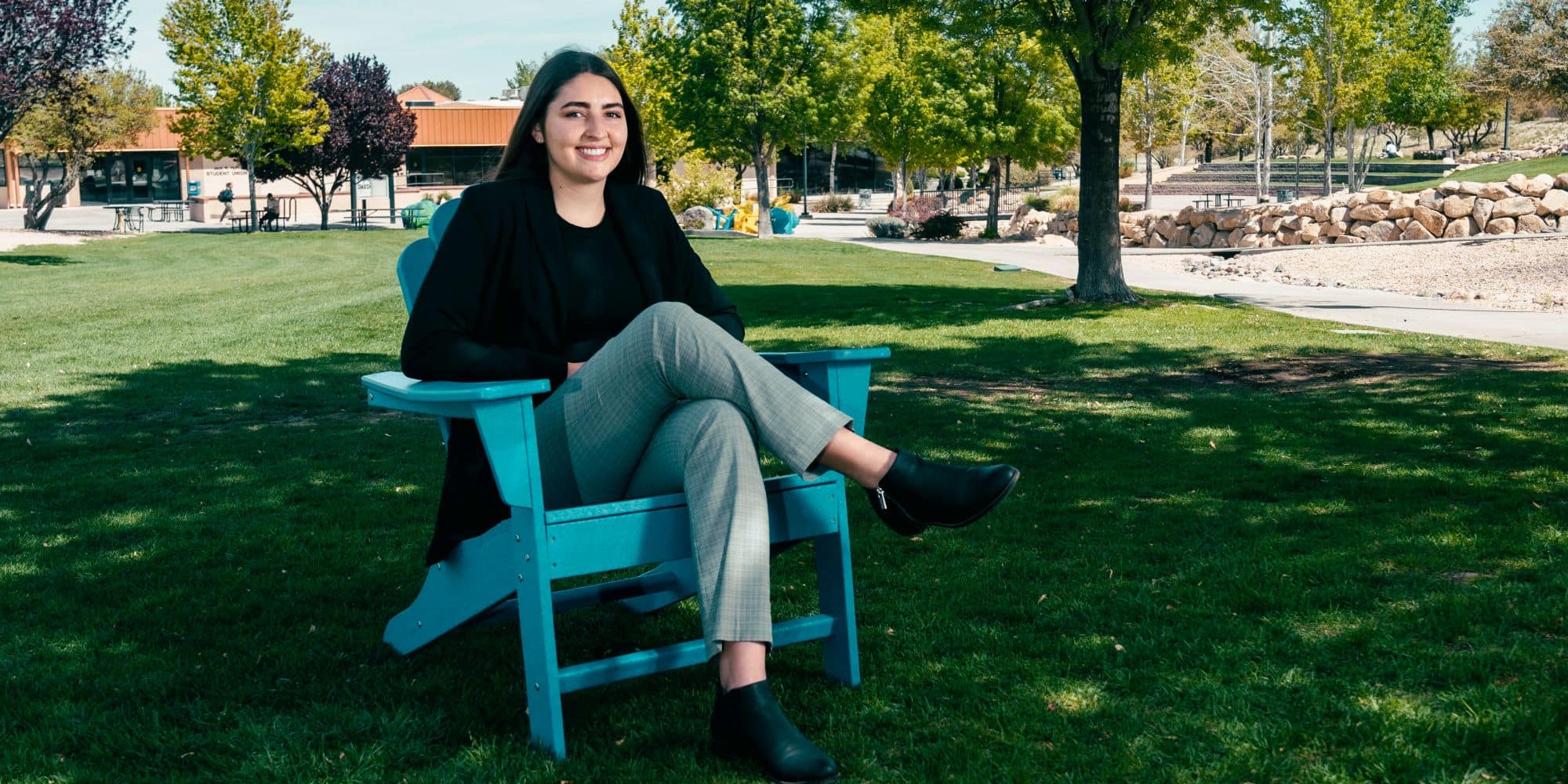 Student seated in Adirondack chair on campus lawn with legs crossed, trees and landscaped walkways around a low academic building in background.