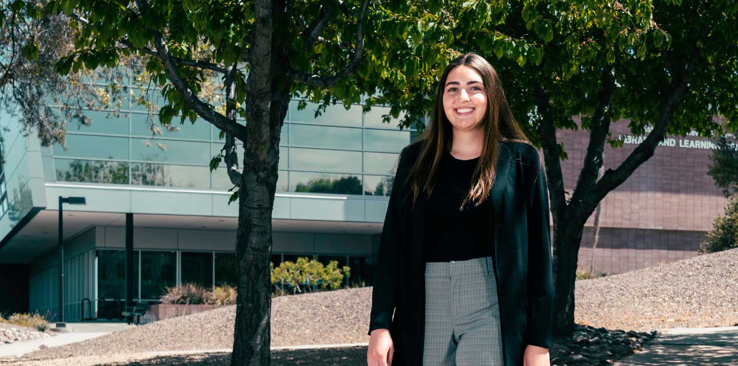 Student standing outdoors under trees in front of a modern campus building with large glass windows and brick facade, landscaped grounds with gravel and shrubs, smiling at camera.