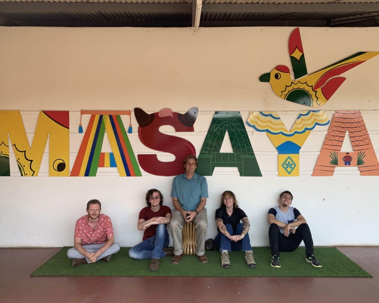 Five people dressed casually pose under a colorful mural with a folk art bird and the word, Masaya.