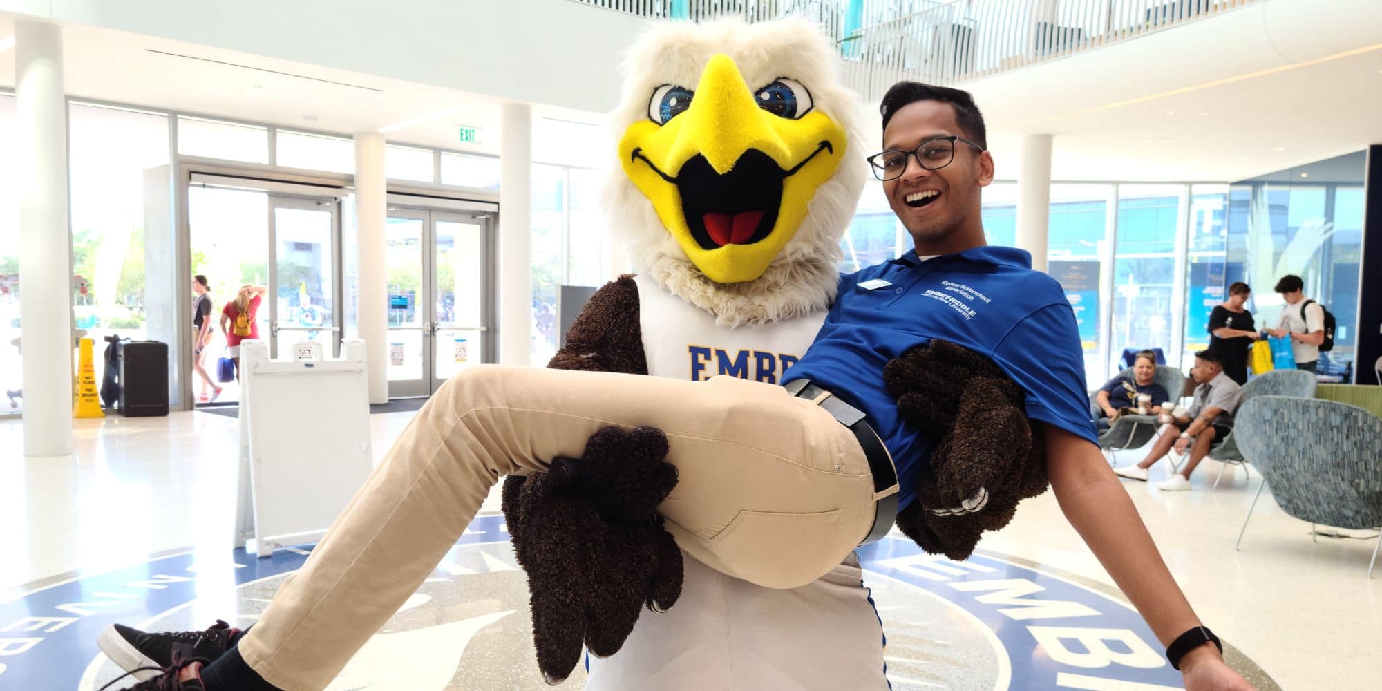 Embry-Riddle eagle mascot holds a smiling student in his wings, in a lobby with glass doors, seating areas, and campus branding on the floor.