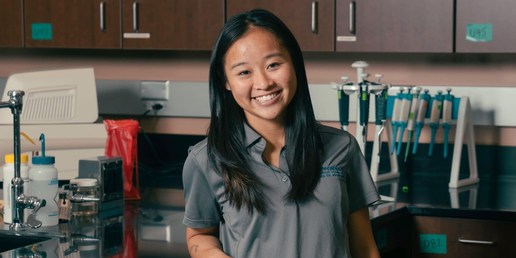 Raelyn, wearing a grey button down shirt with the Embry-Riddle wordmark on the chest, poses smiling in a lab.