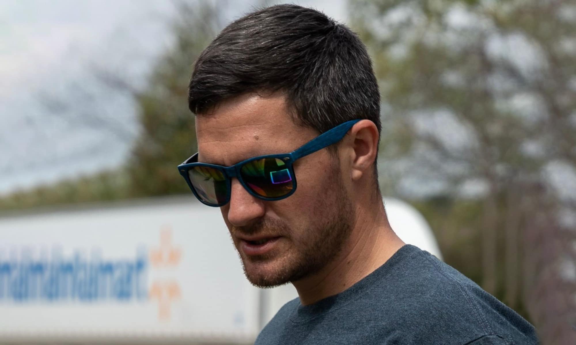 Paul, wearing sunglasses and a grey t-shirt, looks down thoughtfully. He is standing outdoors and a Walmart semi truck is behind him.