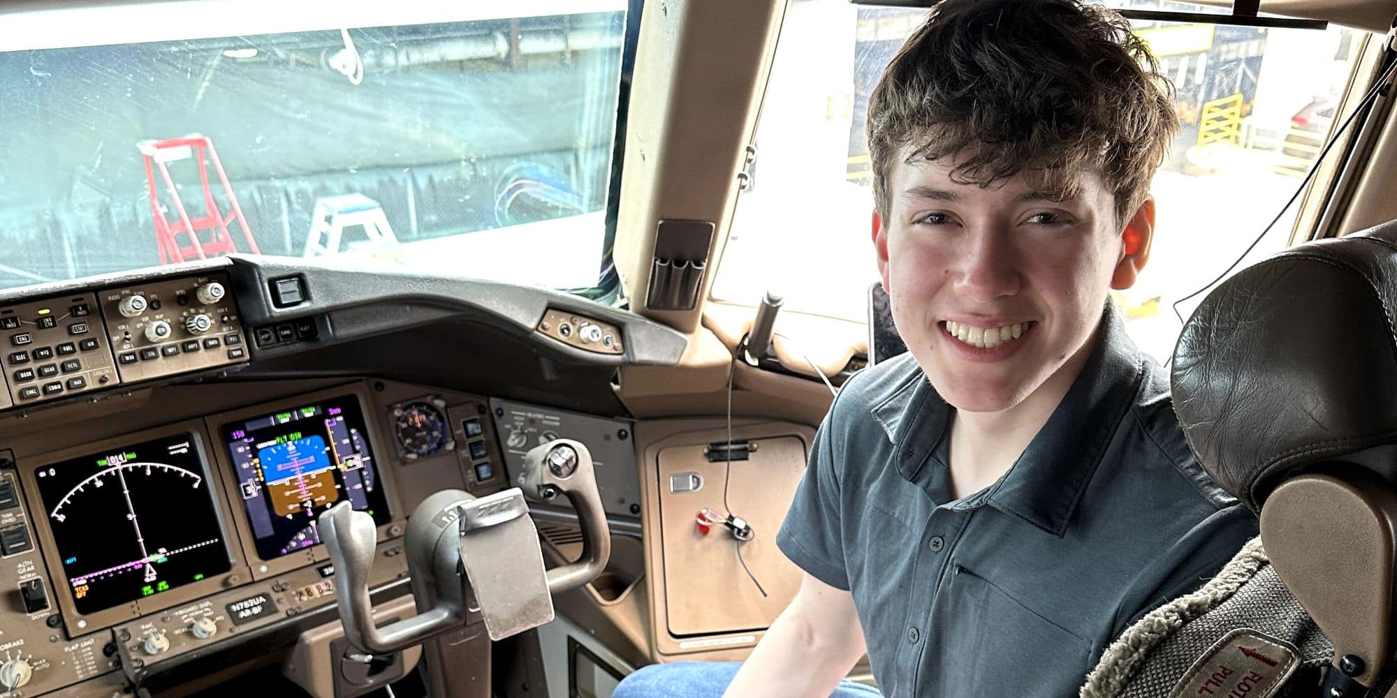 Student smiles at the camera, seated in the glass cockpit of a multi-engine aircraft with avionics displays and flight controls visible.