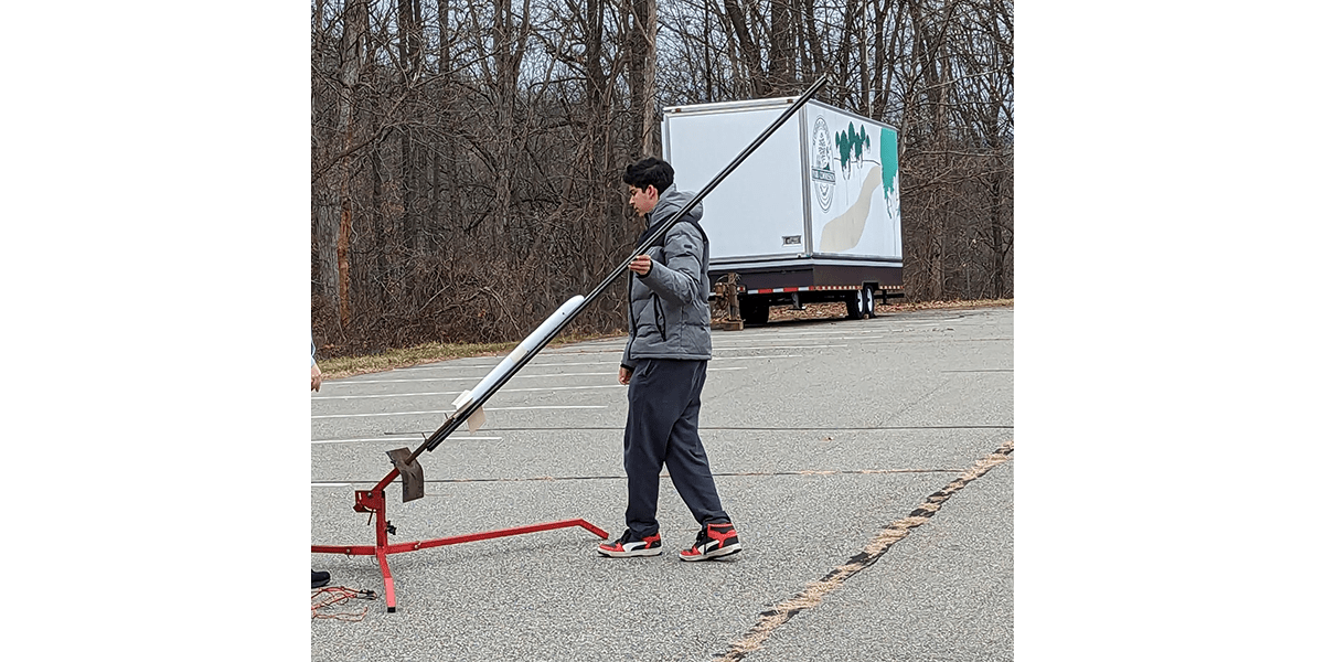 Adam Jain wears a grey puffer jacket and sweat pants as he sets up a rocket on a launch in a parking lot - winter-bare trees are seen in the background.