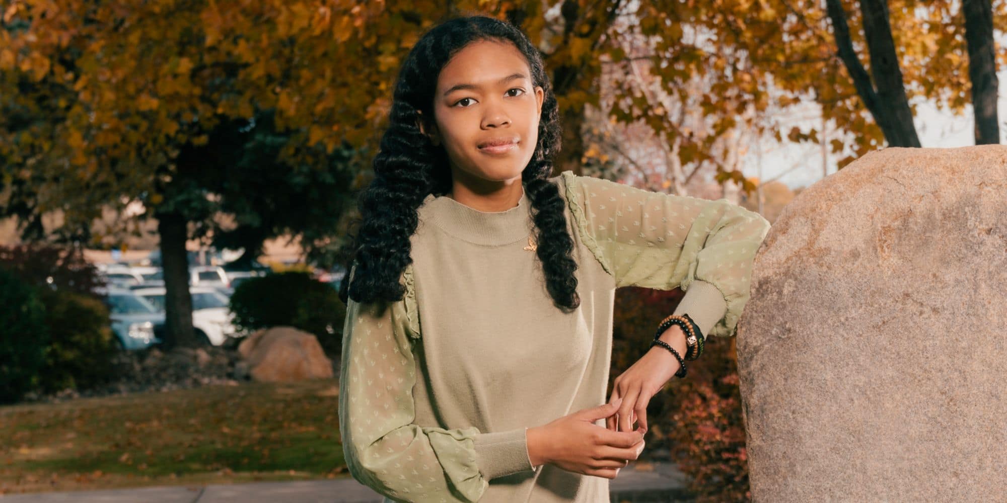 Shii-Ann Madison, wearing a green sweater, leans against a rock, autumn-colored trees in the background.