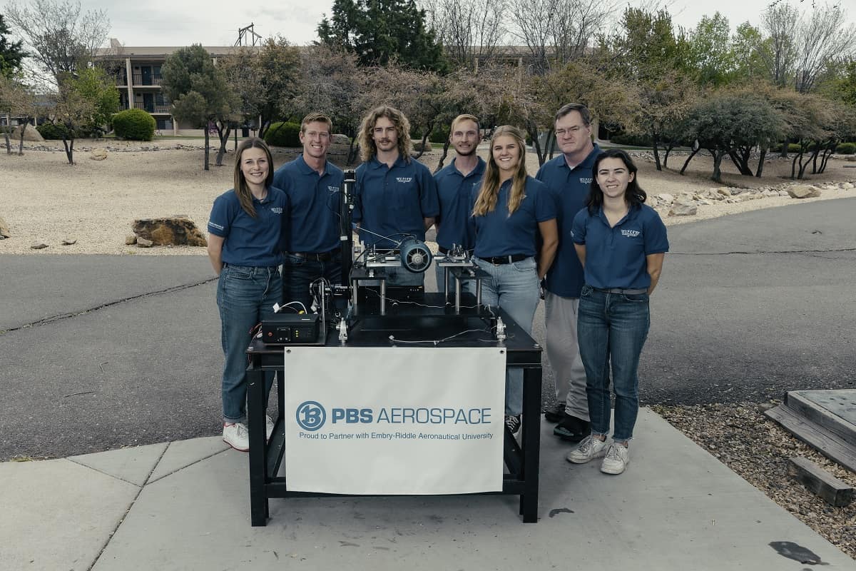 Group of people in blue shirts standing behind table displaying a model of a engine testing facility.
