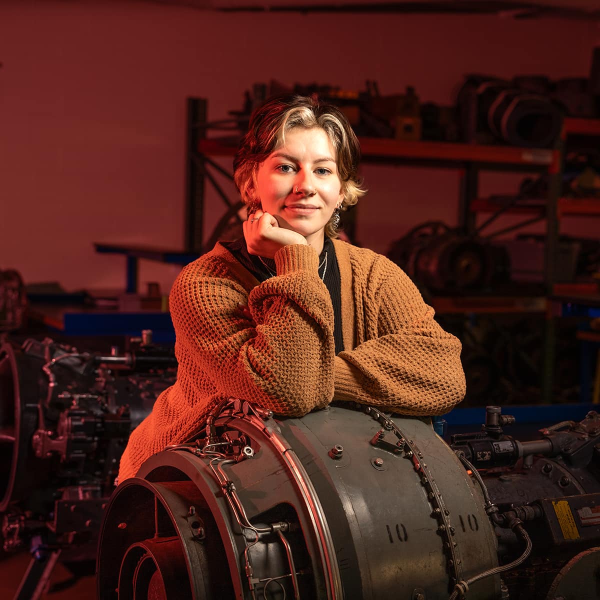 Calen, a woman with light skin tone and short, two-toned hair, poses with her chin in her hand in a maintenance shop.