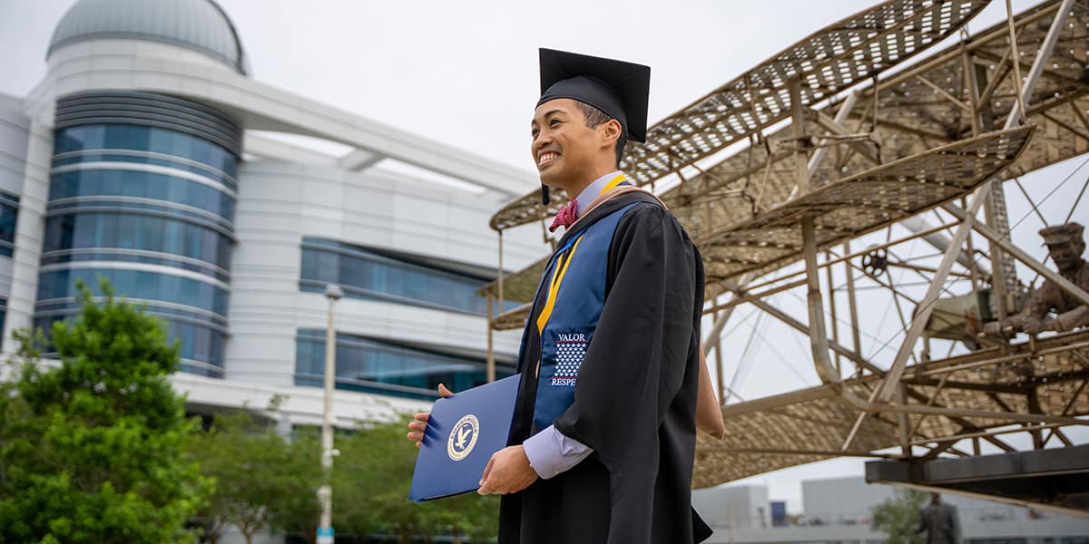 East Asian man wearing graduation cap and gown with a hood and a blue and yellow sash that reads Valor - Respect. He holds up his diploma cover for a photo in front of the historical biplane replica on the Daytona Beach Campus.