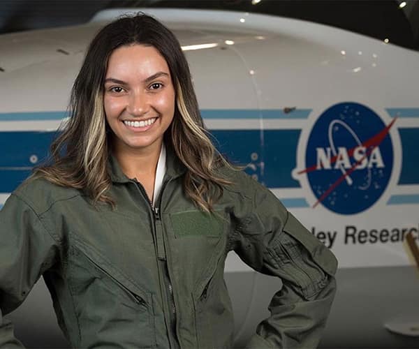 Perla, wearing an army green jumpsuit, poses in front of a NASA research vehicle.