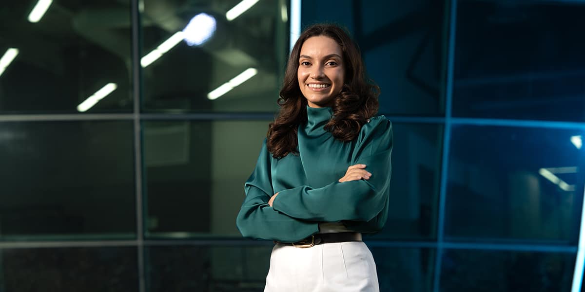 Perla Latorre-Suarez poses in front of a wall of windows; she wears a silky green blouse and white slacks.