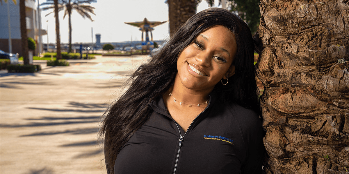 Shyan, a black woman with long hair, poses with a friendly smile, leaning against a palm tree. The Daytona Beach steel abstract plane sculpture, Pathways to the Sky, can be seen in the distance.