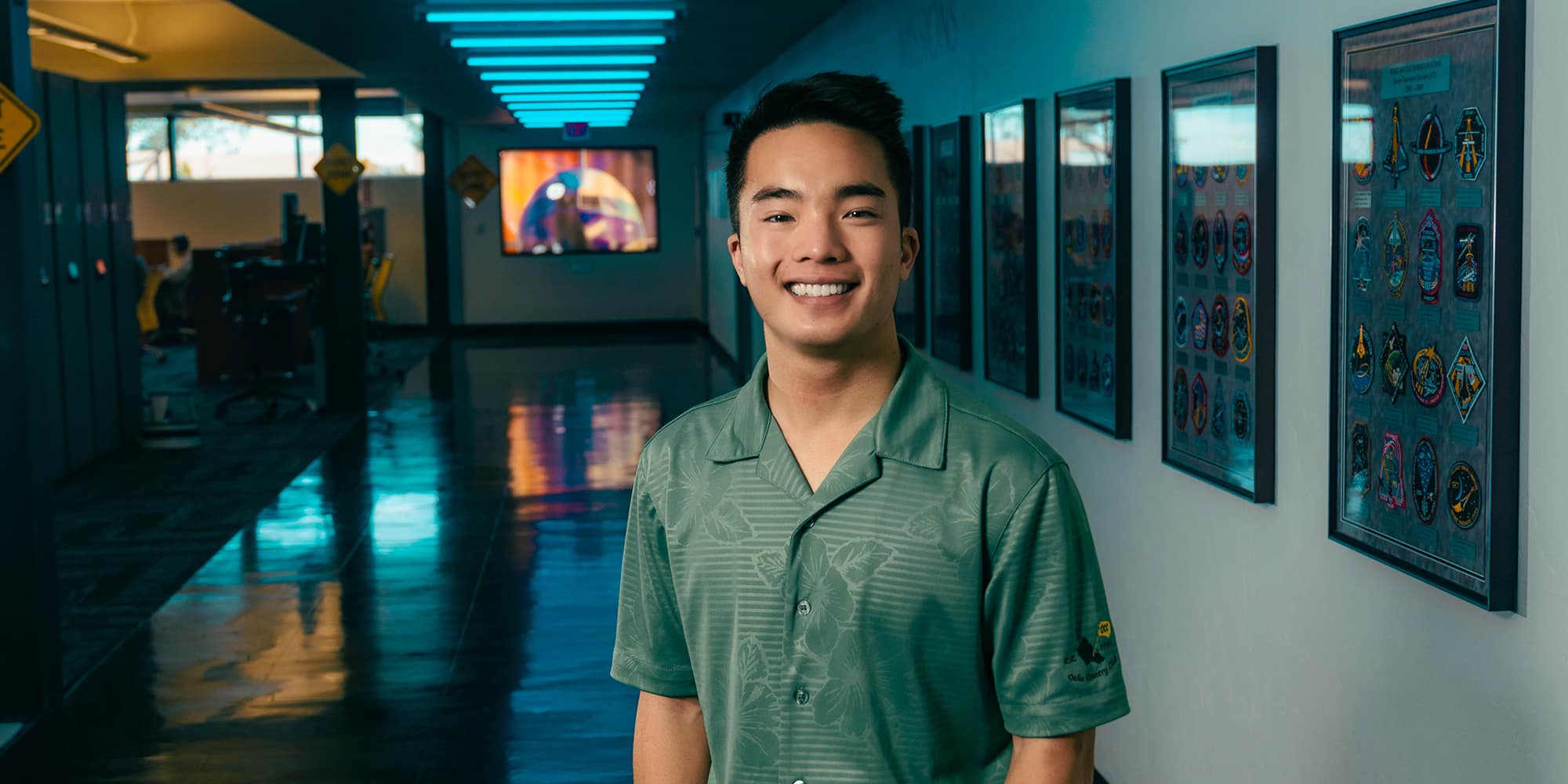 Alexander stands in a darkened hallway lined with framed collections of aviation patches. He has medium skin tone and wears a green shirt with a subtle hibiscus pattern.