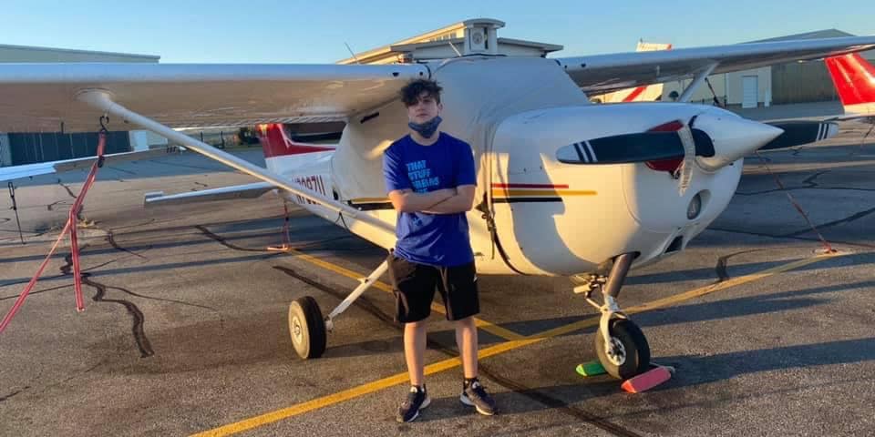 William, a young man with light skin tone and curly hair, poses in a blue t-shirt in front of a small piston aircraft.