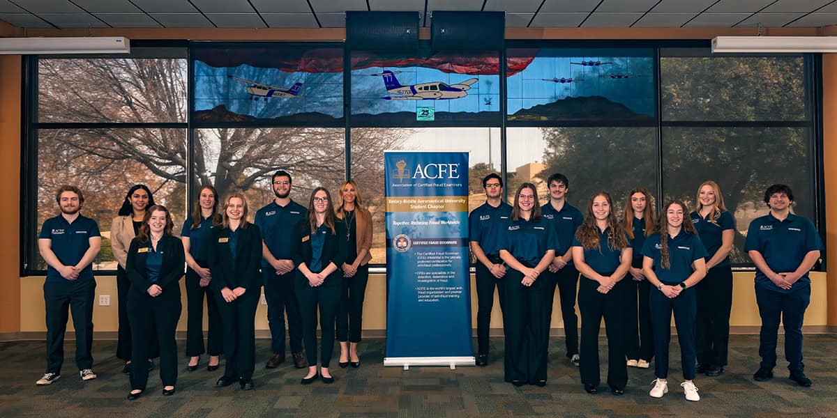 Sixteen men and women - all but two wearing navy polos embroidered with the Embry-Riddle wordmark - stand alike with their hands clasped in front of them. An ACFE banner is set up among them and a stained glass window depicting small planes in flight is set in the window above.