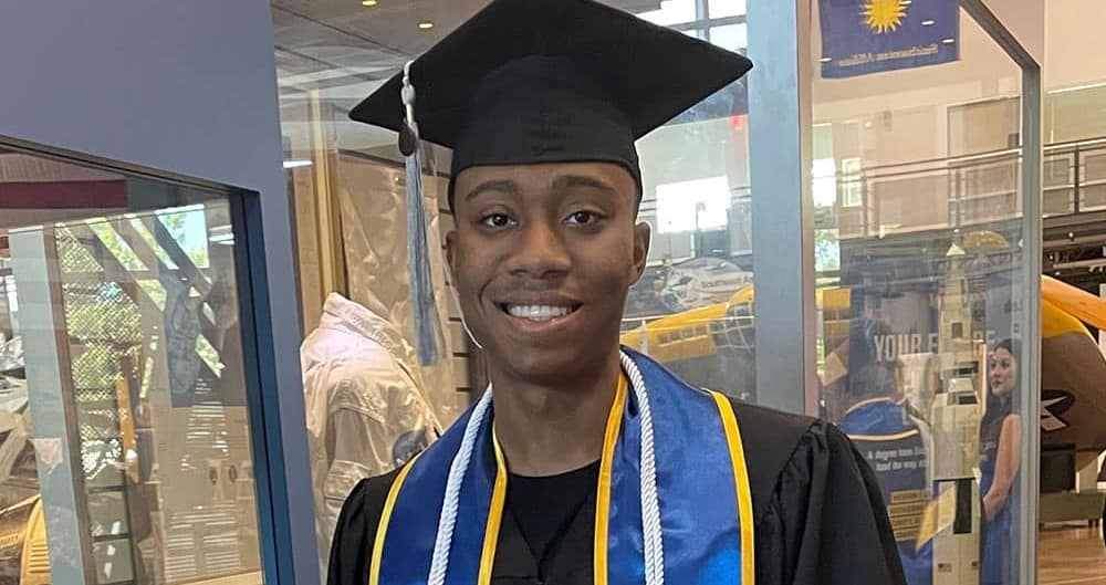 Kegan, a man with dark skin tone, wears a graduation cap and gown, a blue and yellow sash, and white cords.
