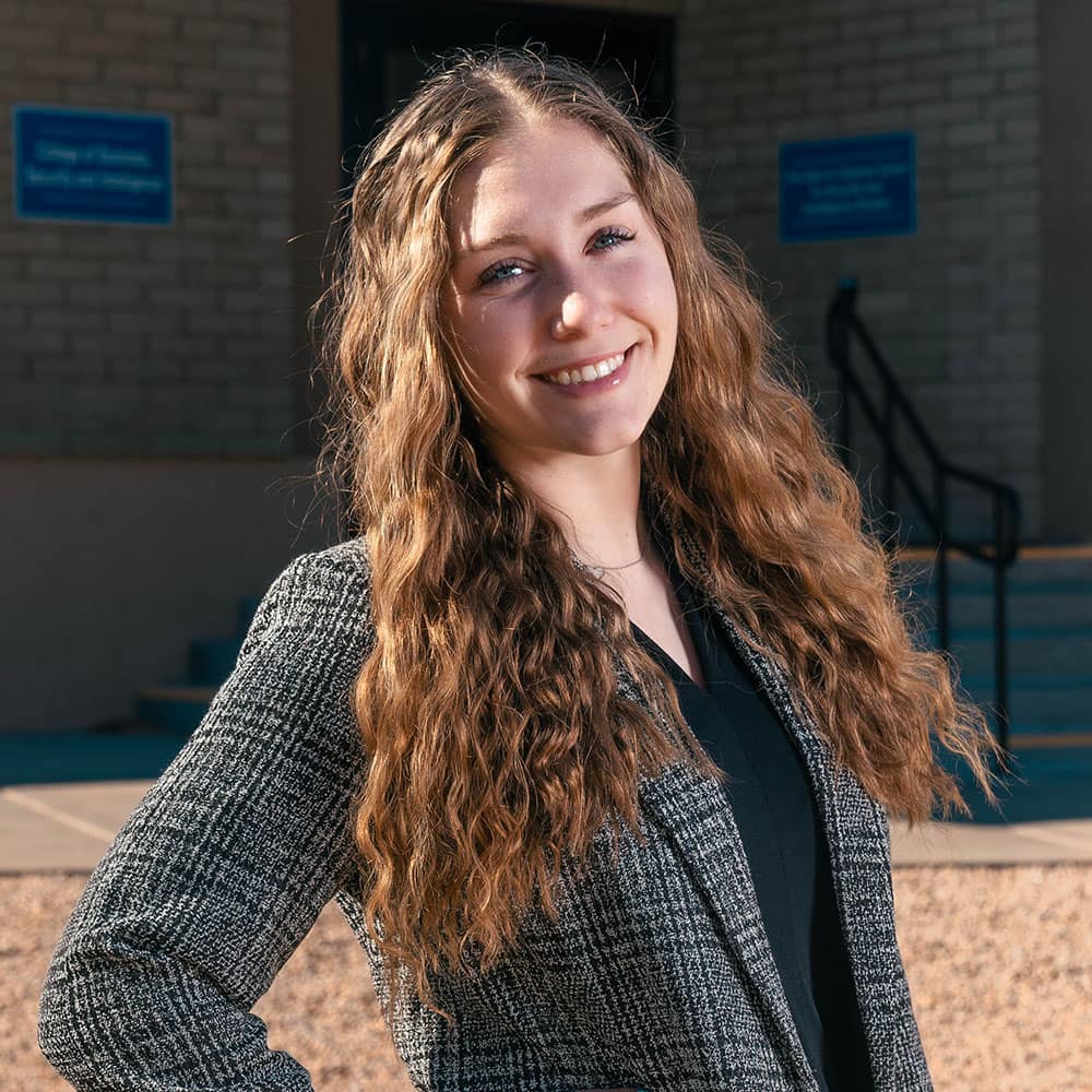 Meredith wears a dark grey suit coat and poses in front of a brick building.