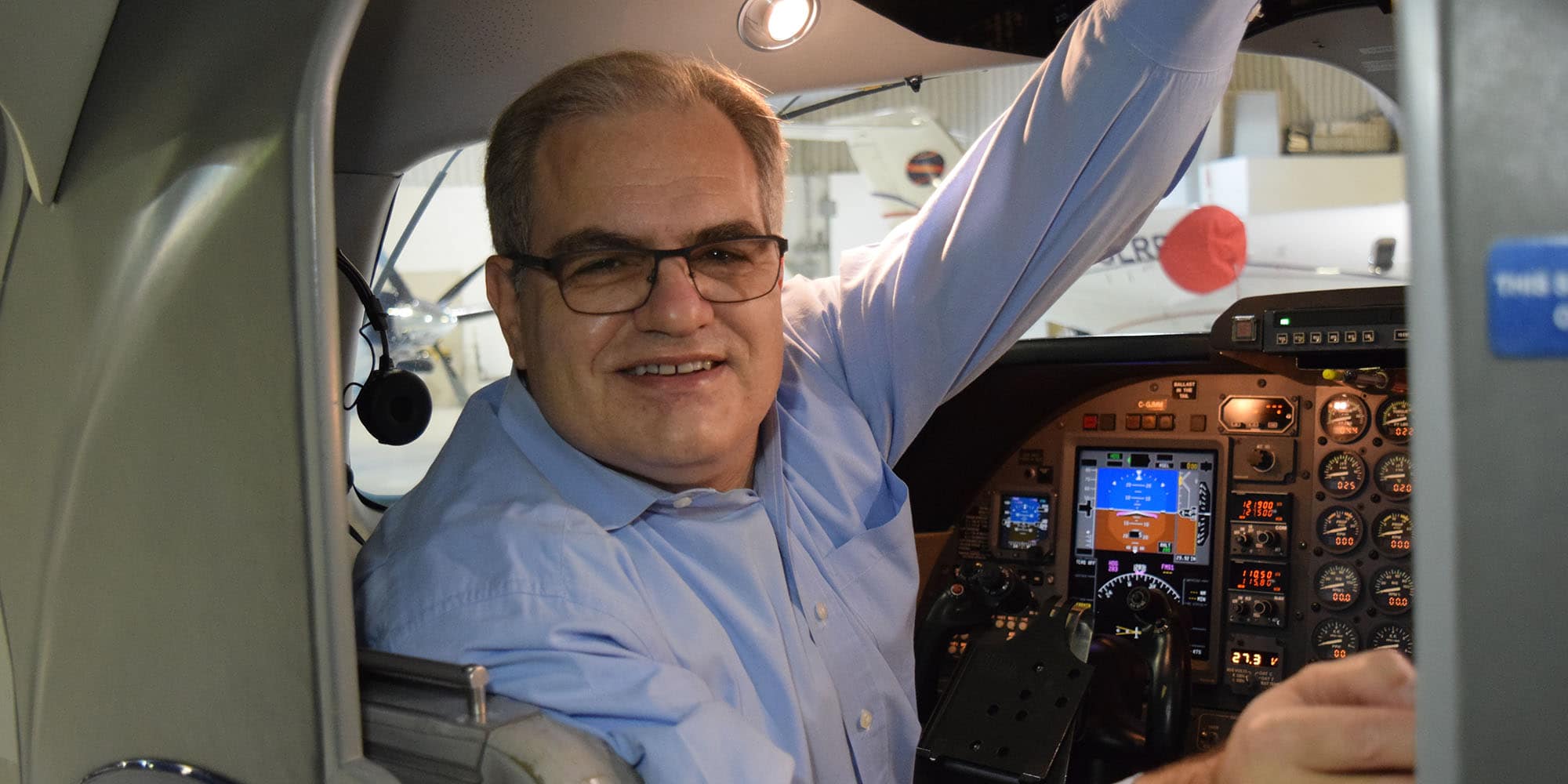 John sits in a small flight deck, wearing a blue button-down shirt.