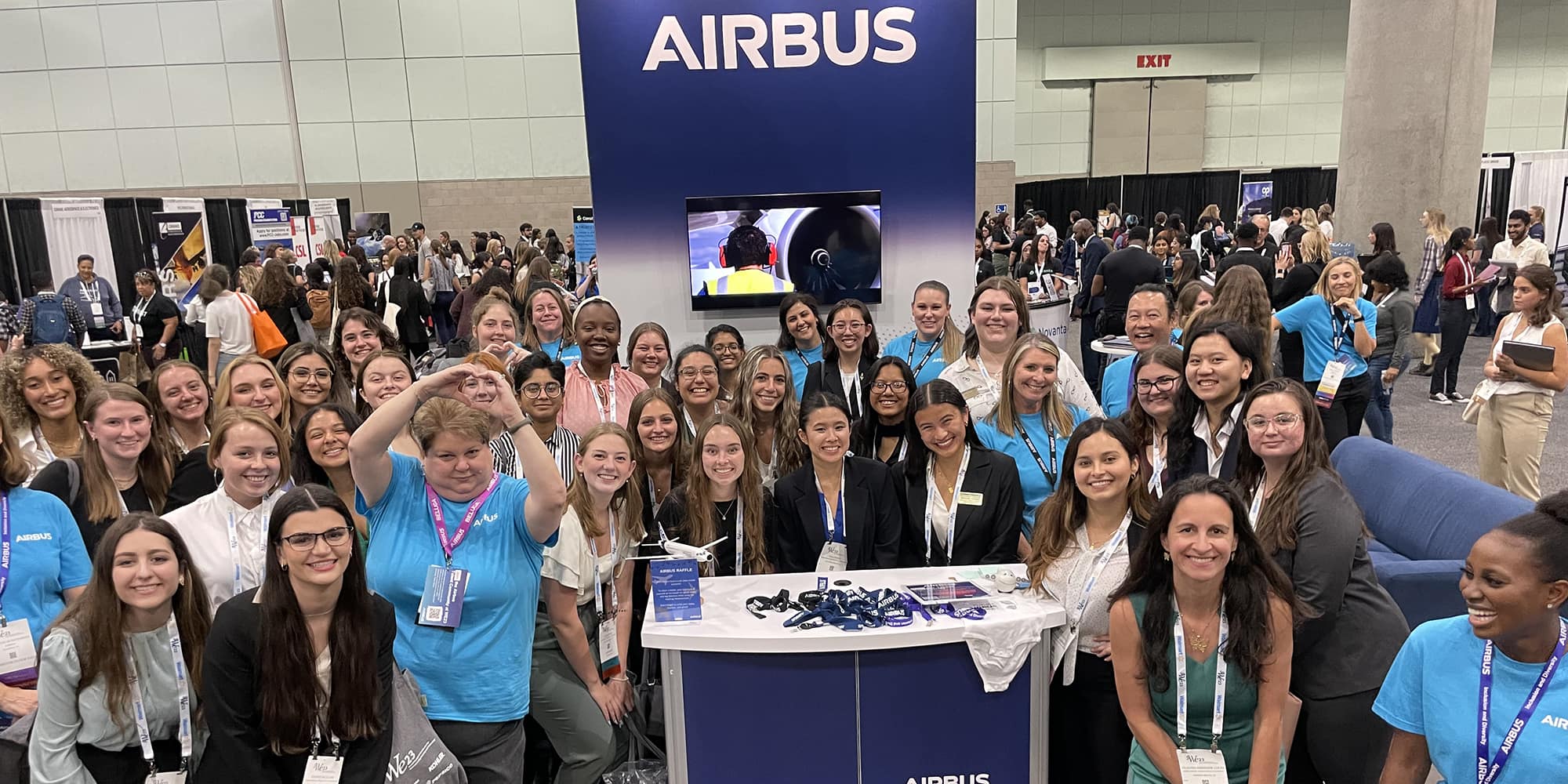 About two dozen, mostly women, pose around the Airbus booth in an expo hall, smiling at the camera. A couple of people hold up a heart sign made with their hands.