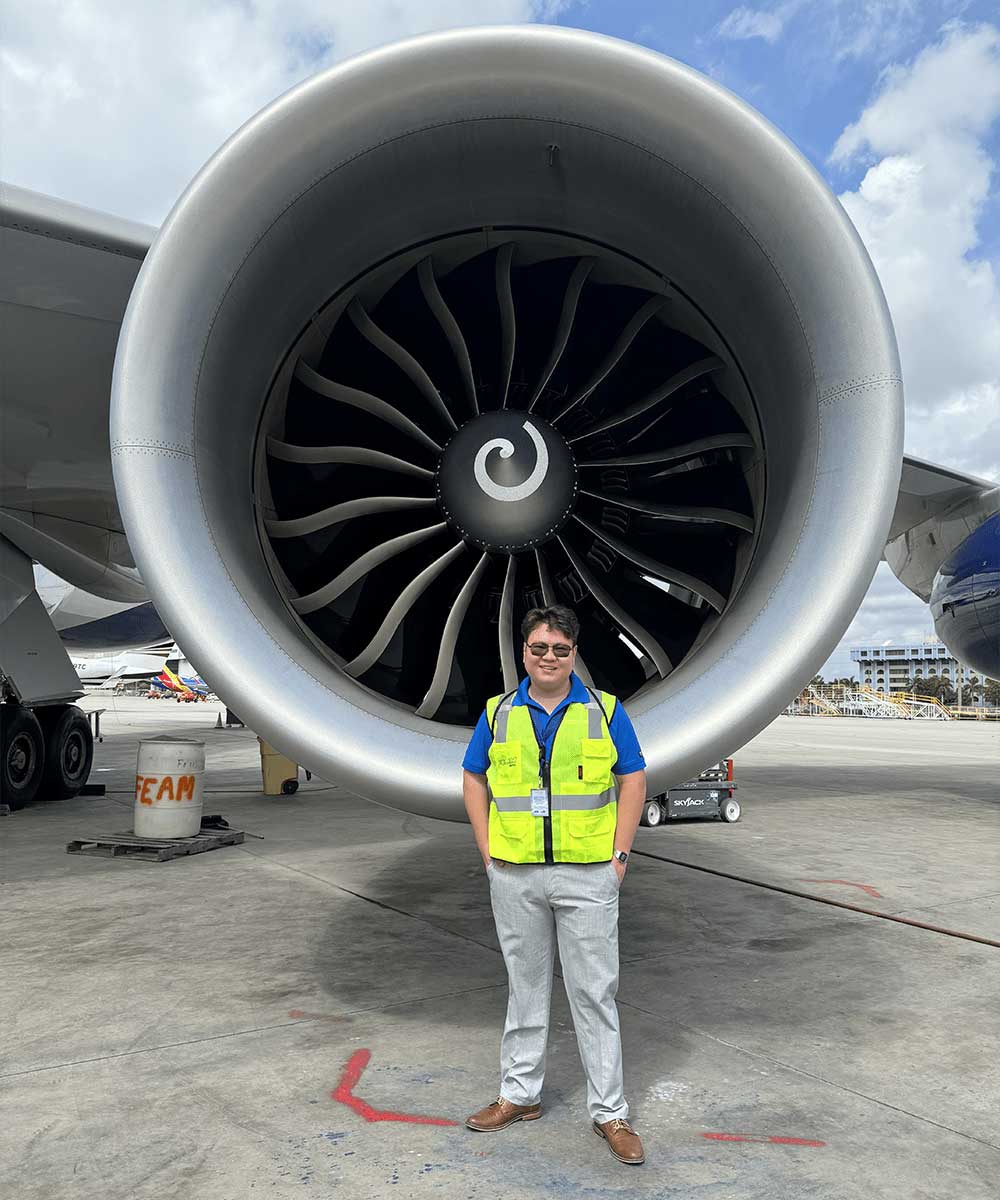 Student in safety vest standing in front of large turbofan engine on airport ramp with fan blades and nacelle visible.