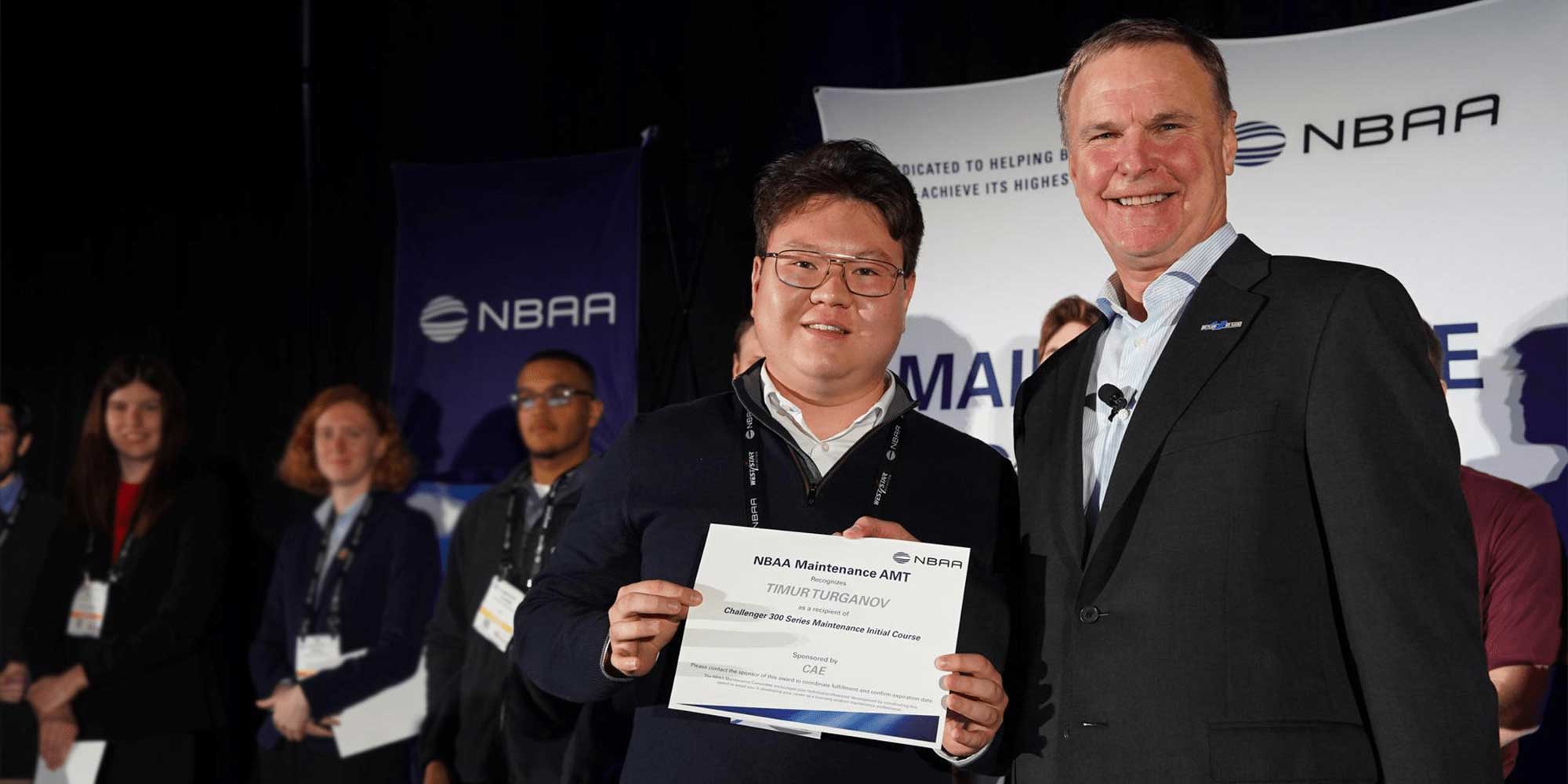 Timur Turganov receiving NBAA Maintenance AMT certificate on stage with presenter and industry event backdrop.