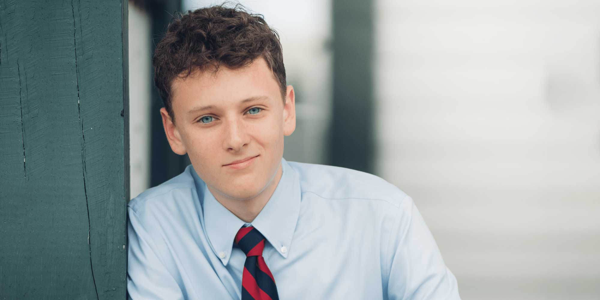 Alexander White leans against a wall, wearing a light blue button-down shirt and a red and blue striped tie.