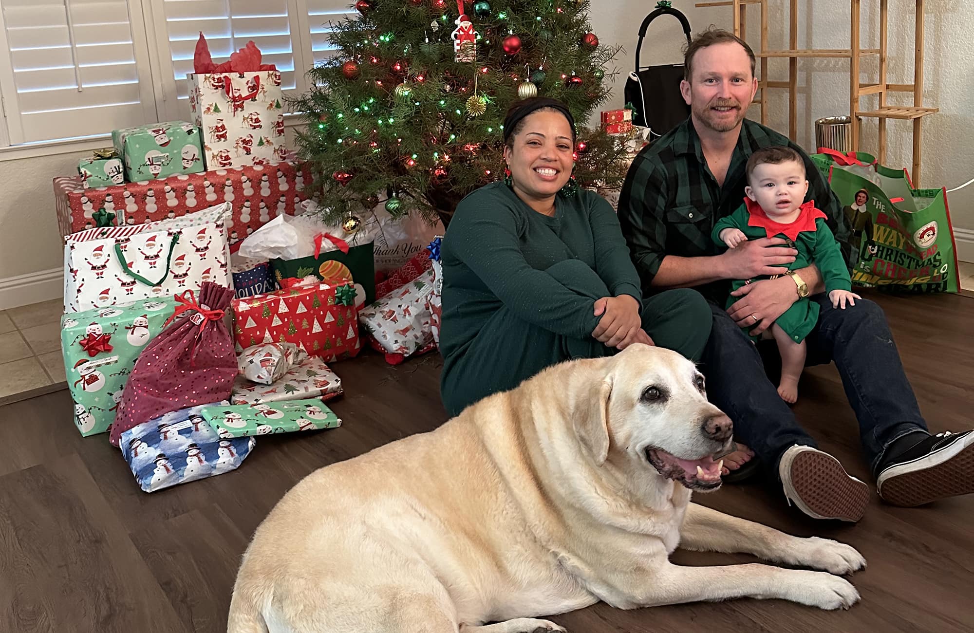 David holds a child in his lap and poses with his wife and a yellow lab in front of a decorated Christmas tree surrounded by wrapped gifts.