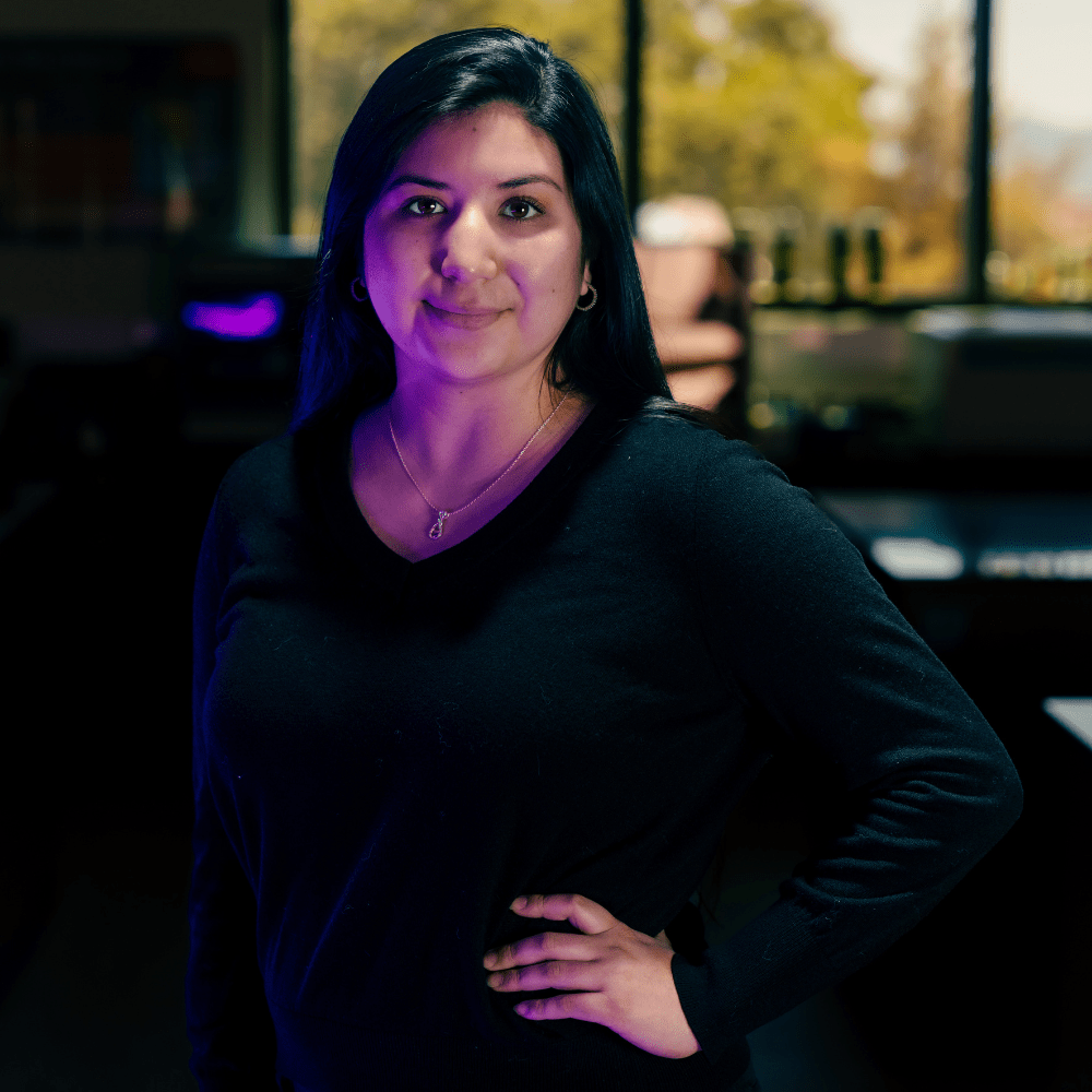 Narayt Salcido poses with a hand on her hip, wearing a dark top and a necklace; windows in the background show a view of greenery.