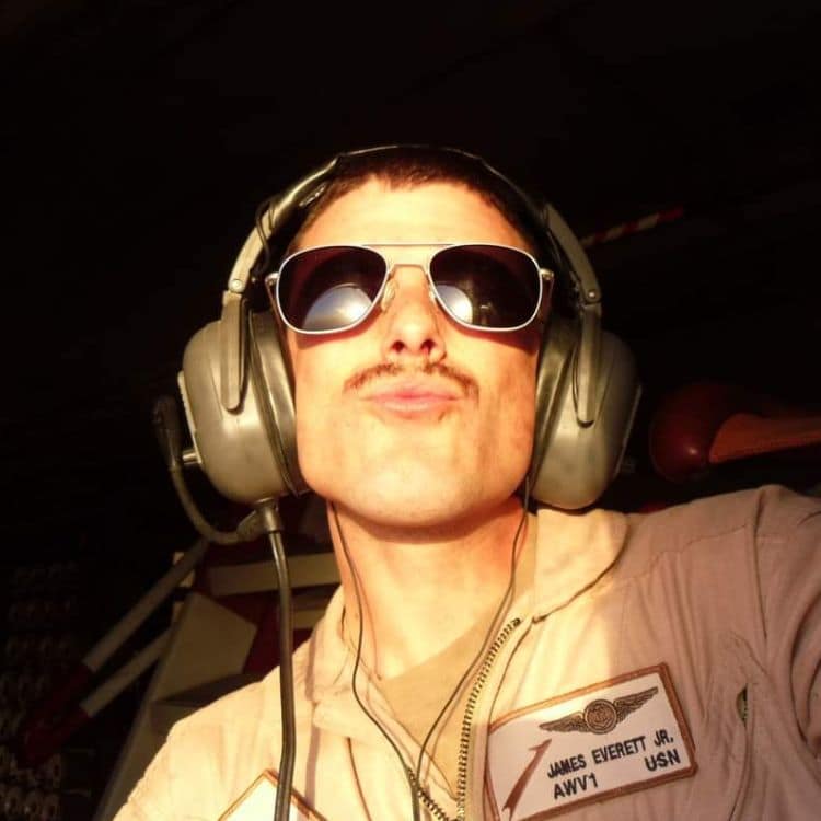 Person wearing aviation headset and aviator sunglasses in flight suit with name patch, posing in low light.