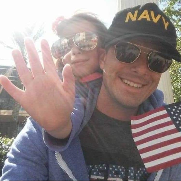 Adult wearing sunglasses and a cap embroidered with 'Navy' carries a child waving and holding small American flag outdoors in bright sunlight.