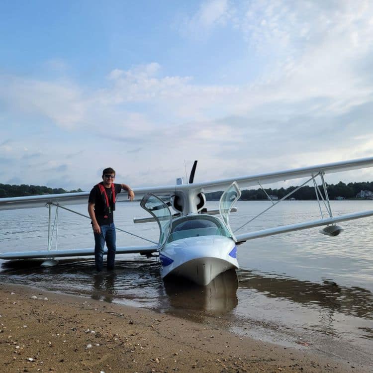 Man stands next to white biplane on shore of lake.
