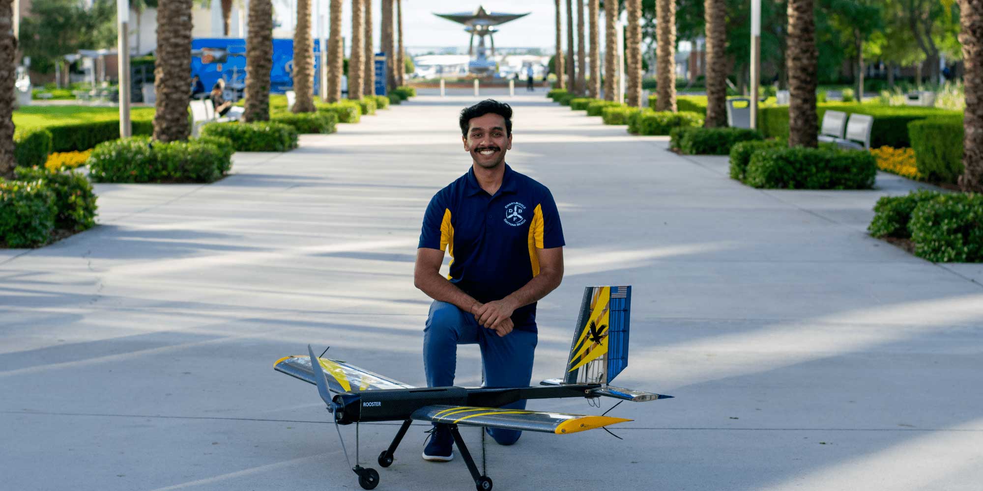Student kneels behind a large fixed-wing radio-controlled aircraft with tricycle landing gear and high-aspect-ratio wings on a campus walkway lined with palm trees.