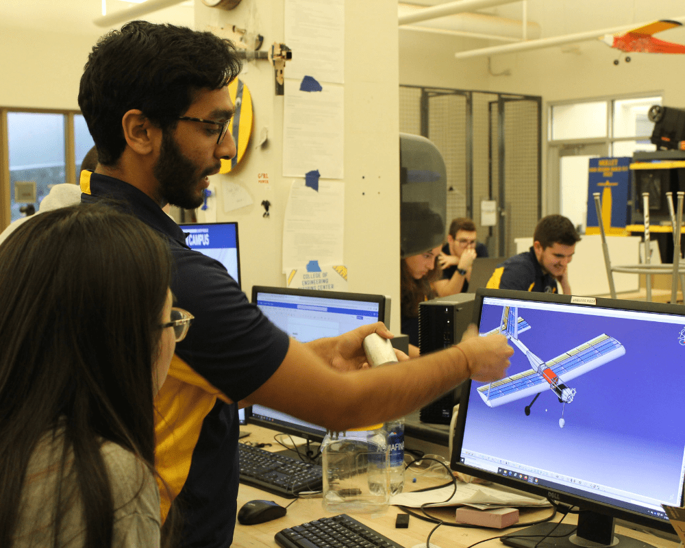 Person demonstrates a CAD model of a fixed-wing aircraft on a computer monitor, pointing at wing and fuselage components while a student observes, aerospace lab with workstations and tools in the background.