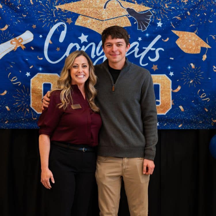 Teacher and student stand together in front of graduation banner.