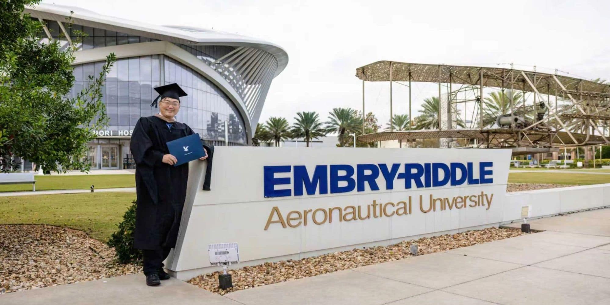 Man in cap and gown stands next to sign stating Embry-Riddle Aeronautical University.