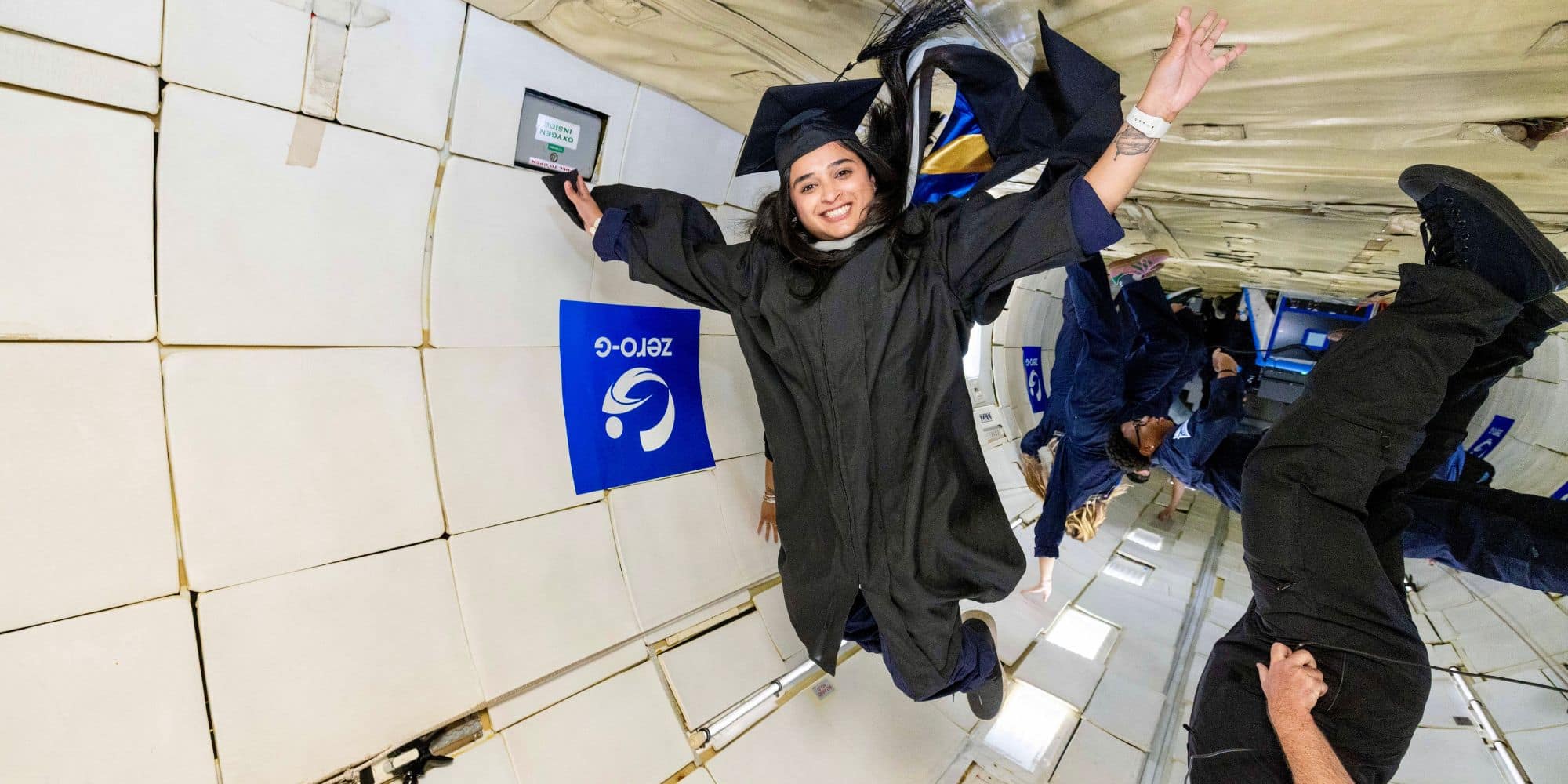 Girl in graduate cap and gown floats inside plane with no gravity.