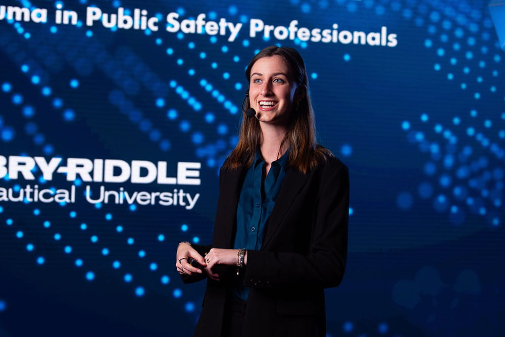 Madison Turner wears a headset and stands on stage in front of an Embry-Riddle background