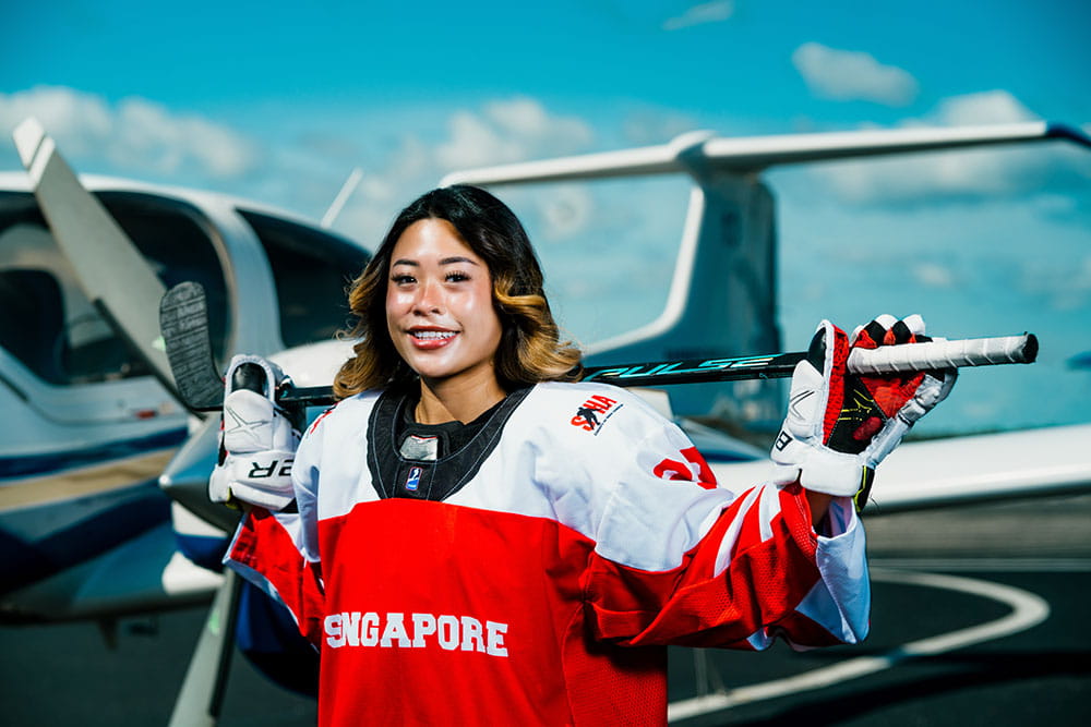 Brianna Tan stands next to a plane while wearing a hockey jersey and holding a hockey stick.