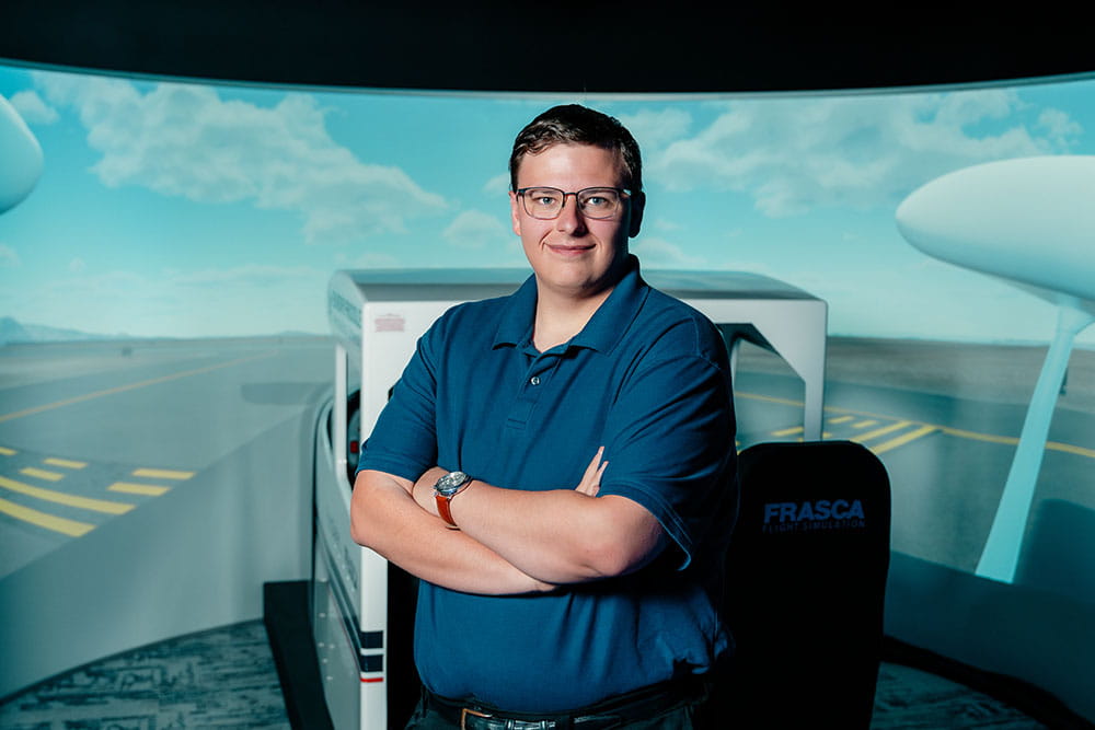 Alec Borneman stands with arms crossed in the Frasca C172S TruFlite AATD Simulator on the Prescott Campus