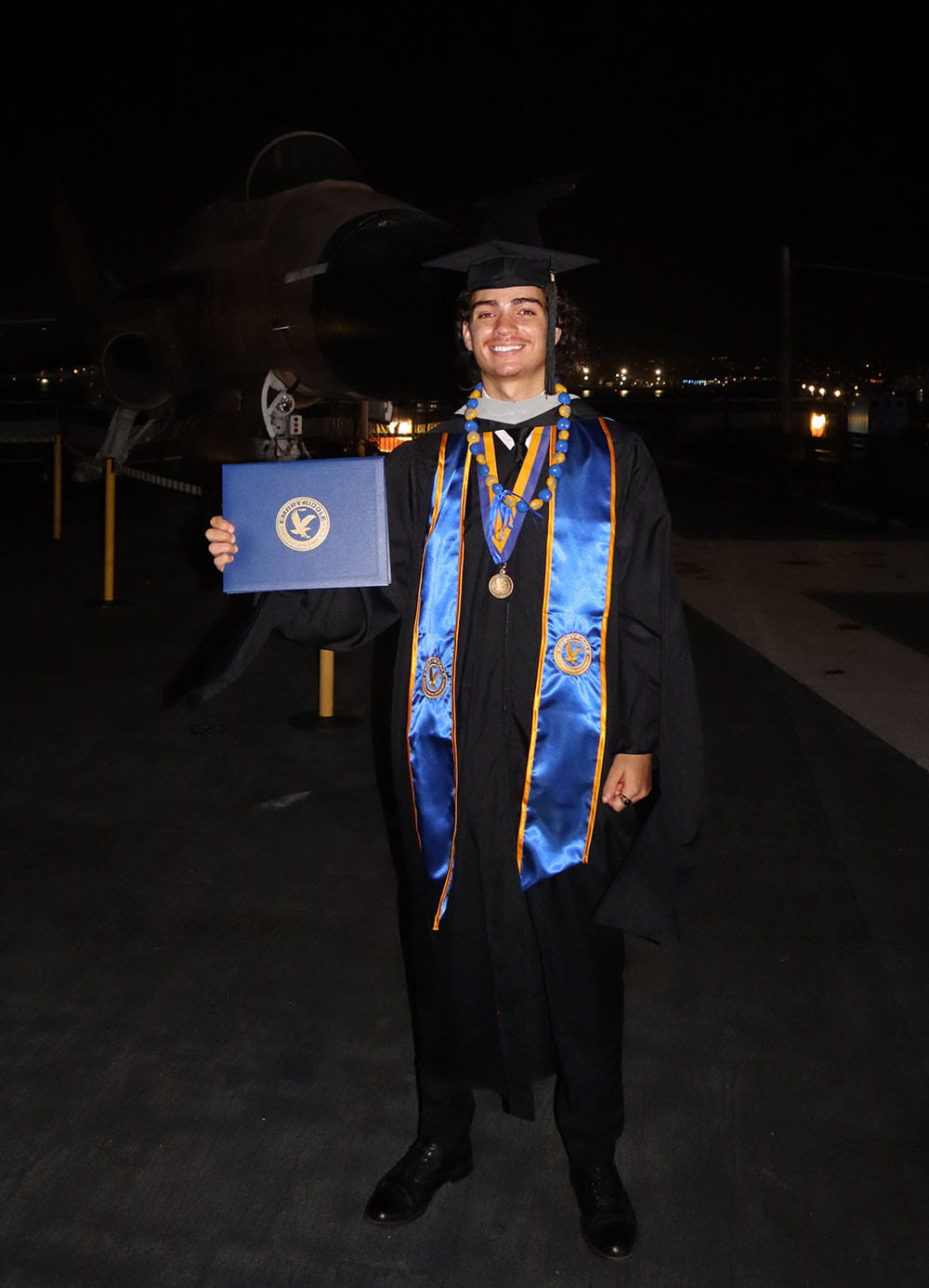 Camryn Huff stands in his graduation cap and gown holding his diploma in front of a plane