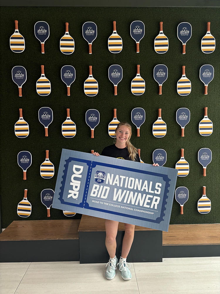 Katie Zint stands in front of pickleball paddles on a wall while holding a large poster that says "Nationals Bid Winner"