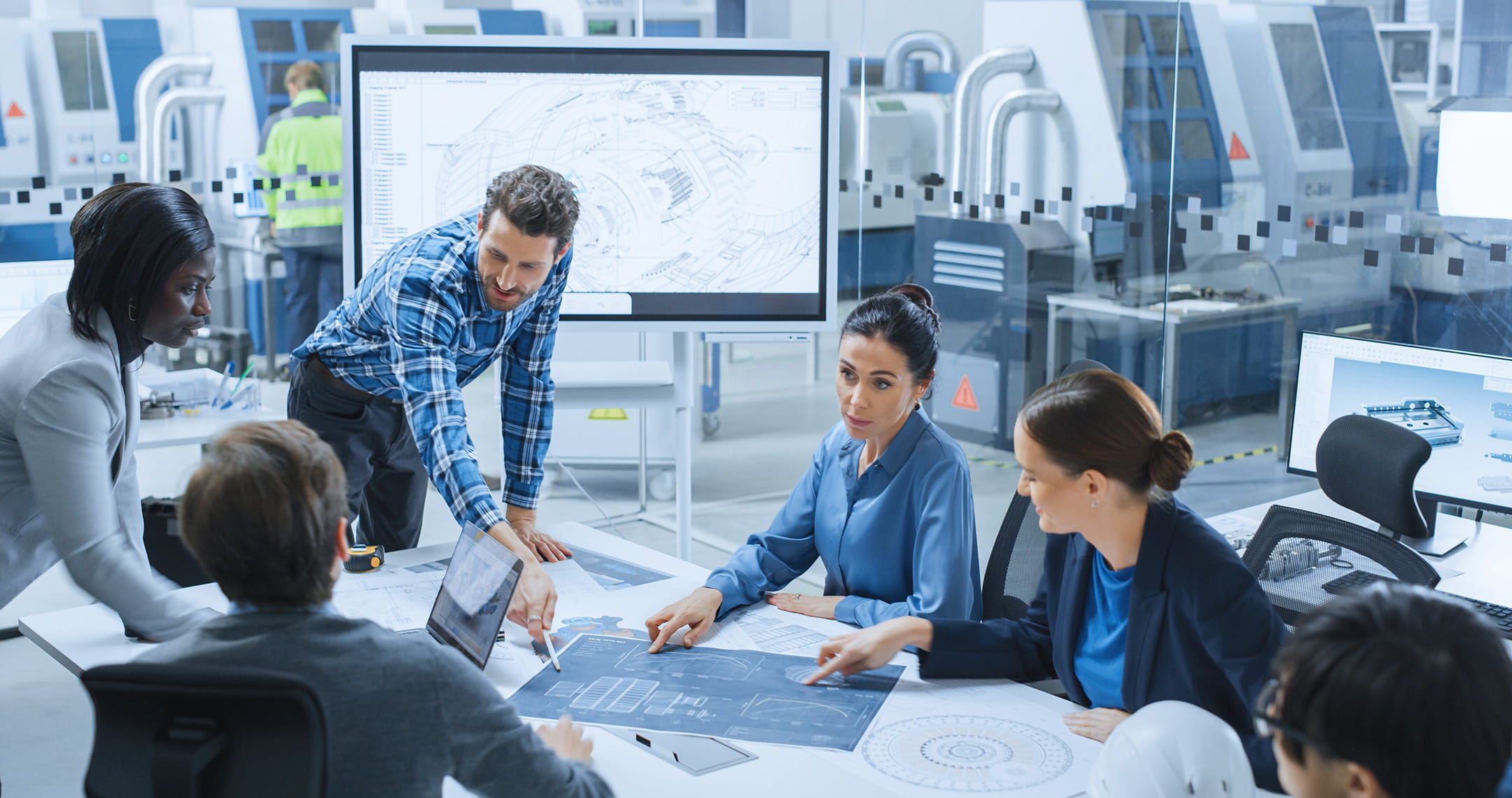 Engineering team reviewing large printed technical schematics at table, pointing to diagrams, CAD model of aircraft component displayed on screen behind them, advanced manufacturing lab with industrial machines in background.